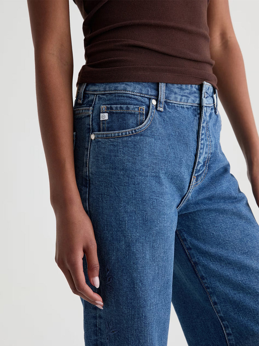 Person wearing a sleeveless brown top and AG Jeans Saige Wide Leg Crop in Malbec blue denim, shown from shoulders to mid-thigh, with one arm relaxed at their side.
