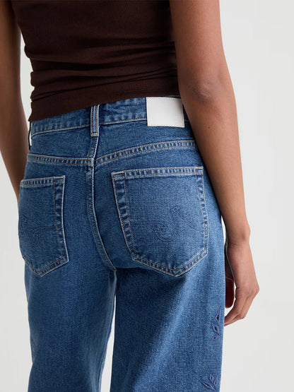 A person is shown from the back wearing AG Jeans Saige Wide Leg Crop in Malbec and a brown ribbed tank top, featuring a white waistband patch with a subtle geometric floral design.