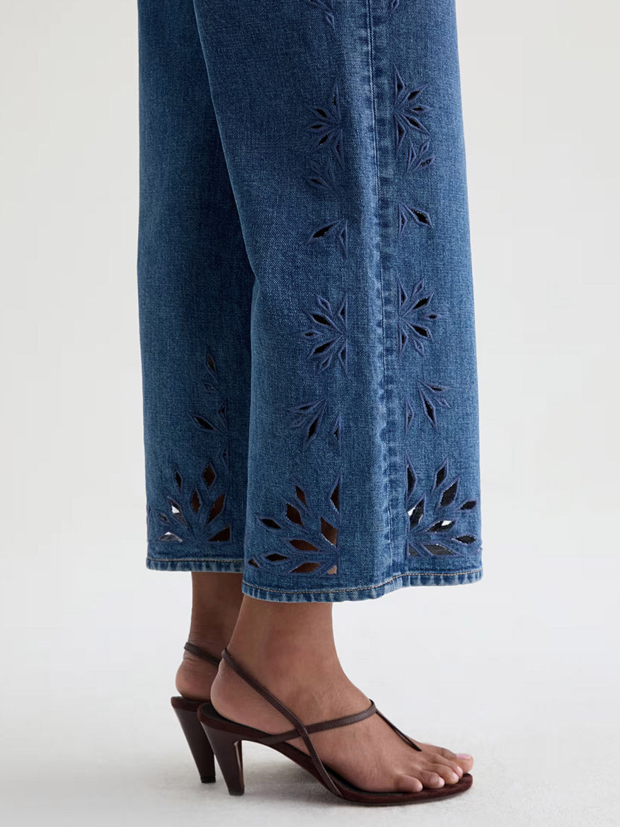 Wearing AG Jeans Saige Wide Leg Crop in Malbec, featuring a geometric floral design near the hem, paired with brown strappy heeled sandals, against a neutral background.