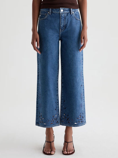 Model wears AG Jeans Saige Wide Leg Crop in Malbec, featuring a geometric floral laser-cut hem, paired with a brown top and heeled sandals. Crafted from Premium Left Hand Denim for a chic look, set against a plain background.