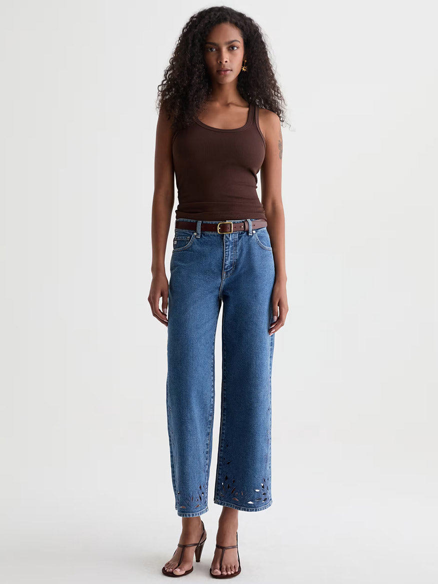 A woman stands against a plain background wearing a brown tank top, AG Jeans Saige Wide Leg Crop in Malbec with a belt, and brown heeled sandals.