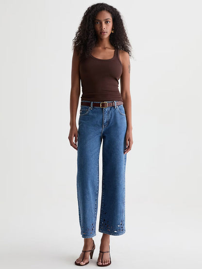 A woman stands against a plain background wearing a brown tank top, AG Jeans Saige Wide Leg Crop in Malbec with a belt, and brown heeled sandals.