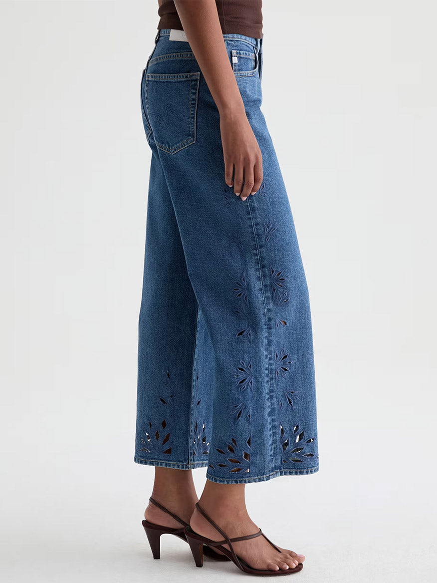 Wearing AG Jeans Saige Wide Leg Crop in Malbec with a geometric floral hem design, a brown top, and dark heeled sandals, a person stands against a plain background.