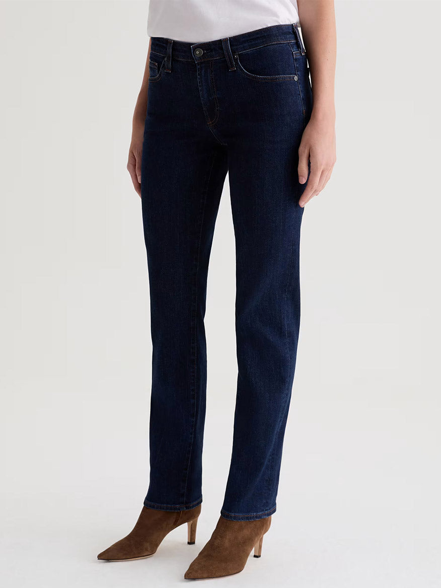 A person wearing AG Jeans Sandra Low-Rise Straight in Modern Indigo, a white top, and brown suede ankle boots stands against a plain light background.