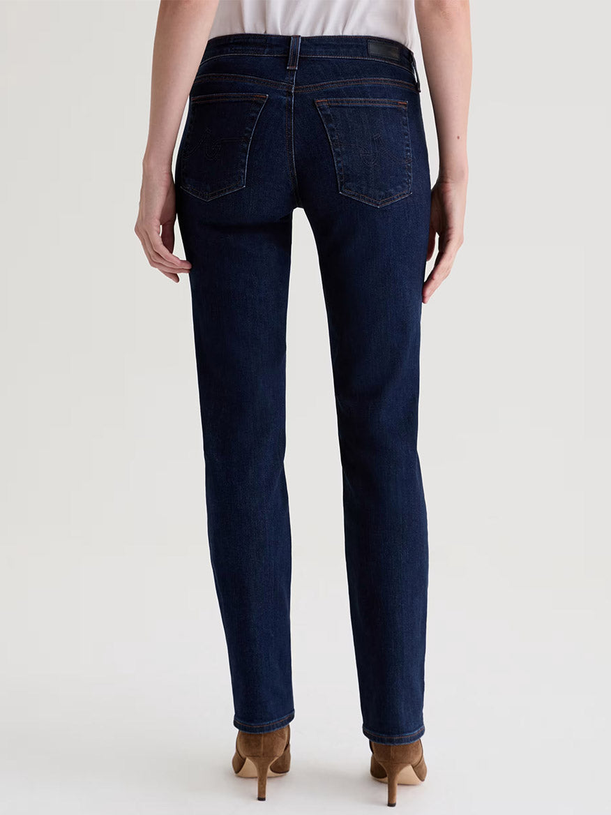 A person shown from the back against a plain light background wears AG Jeans Sandra Low-Rise Straight in Modern Indigo with brown heeled shoes.