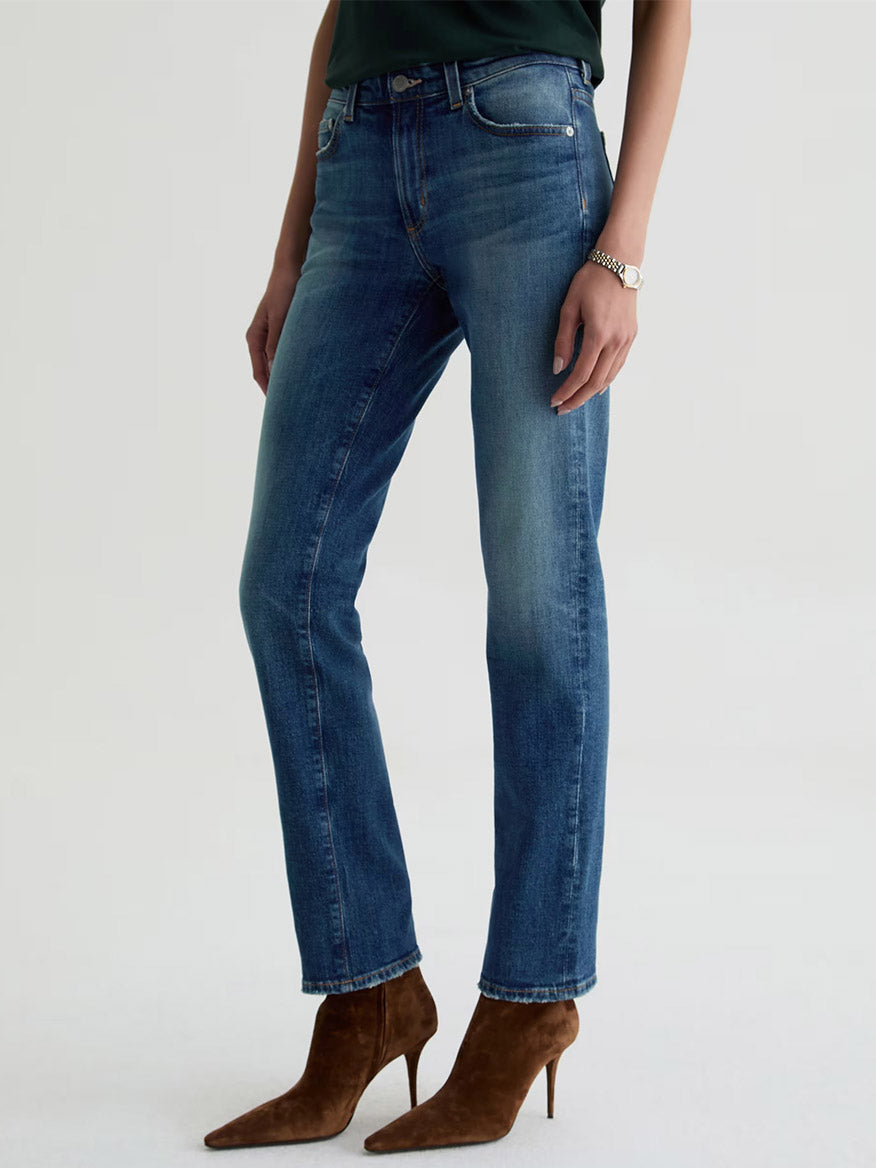 Wearing AG Jeans Sandra Low-Rise Straight in 10 Years Illiad, a dark top, a bracelet, and brown heeled ankle boots, the person stands against a plain light background.