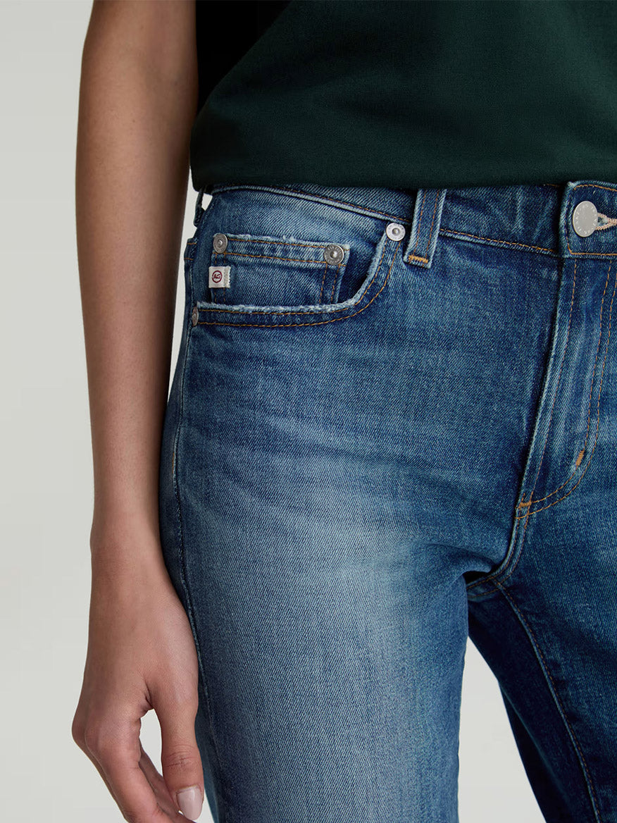 A person in AG Jeans Sandra Low-Rise Straight in 10 Years Illiad, a dark blue wash, and a dark green top, stands with their left hand relaxed at their side, shown from waist to mid-thigh.