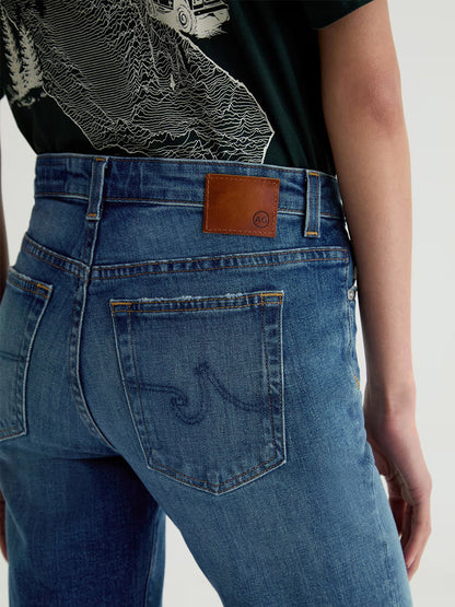 Close-up of a person wearing AG Jeans Sandra Low-Rise Straight in 10 Years Illiad, featuring dark blue wash, straight-leg fit, brown leather patch and wave stitching on the back pocket, paired with a dark shirt with white graphic.