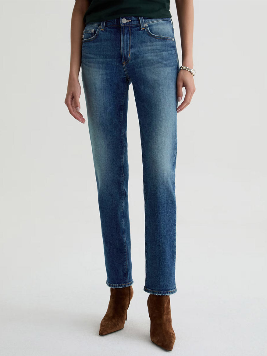 A person wearing AG Jeans Sandra Low-Rise Straight in 10 Years Illiad, a dark top, a bracelet, and brown suede ankle boots stands against a plain light background.