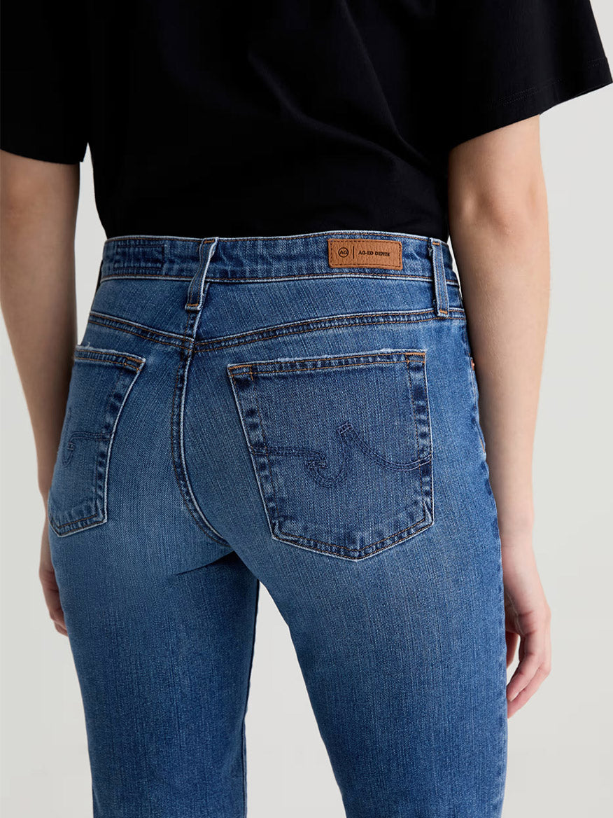 Person wearing AG Jeans Sandra AG-ed™ Low-Rise Straight in 15 Years Prague and a black shirt, shown from the back, with visible back pockets and a brown leather patch on the waistband.