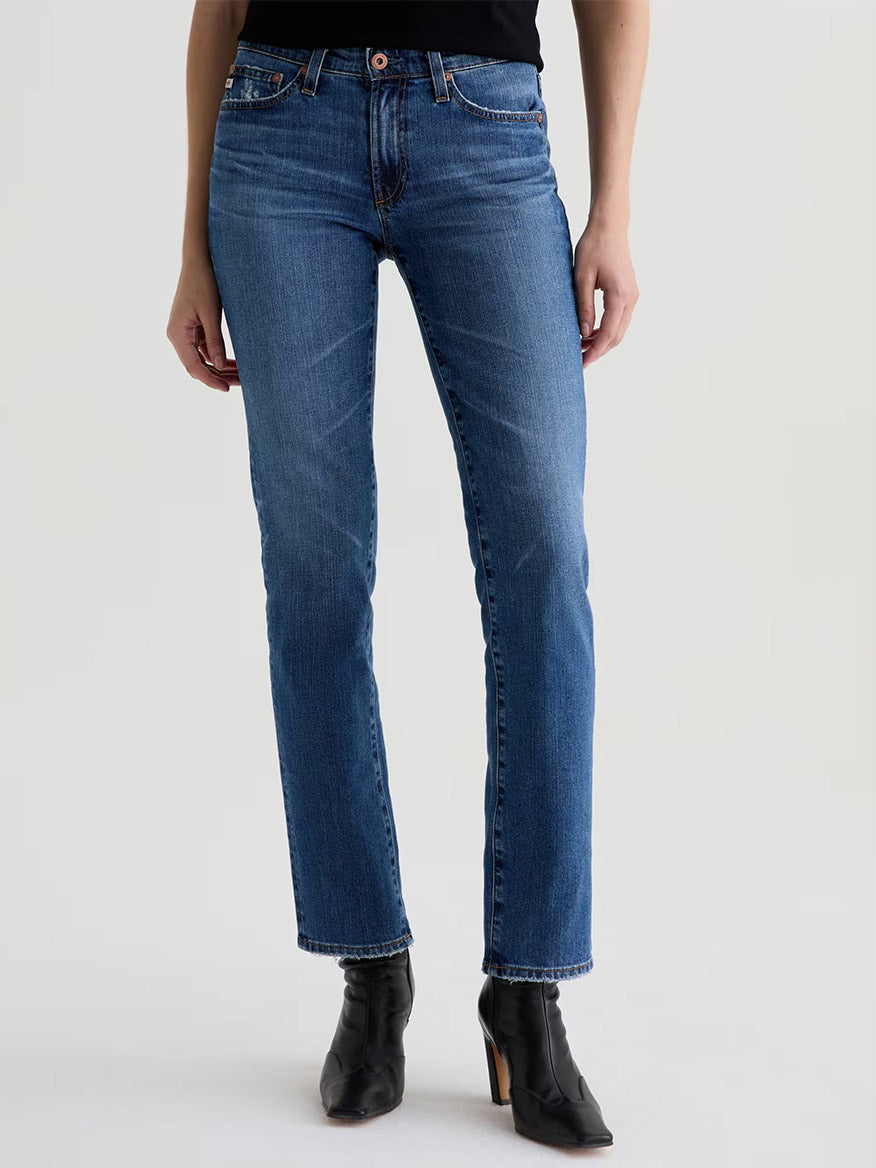 A person wearing AG Jeans Sandra AG-ed™ Low-Rise Straight in 15 Years Prague and black heeled ankle boots stands against a plain light background.