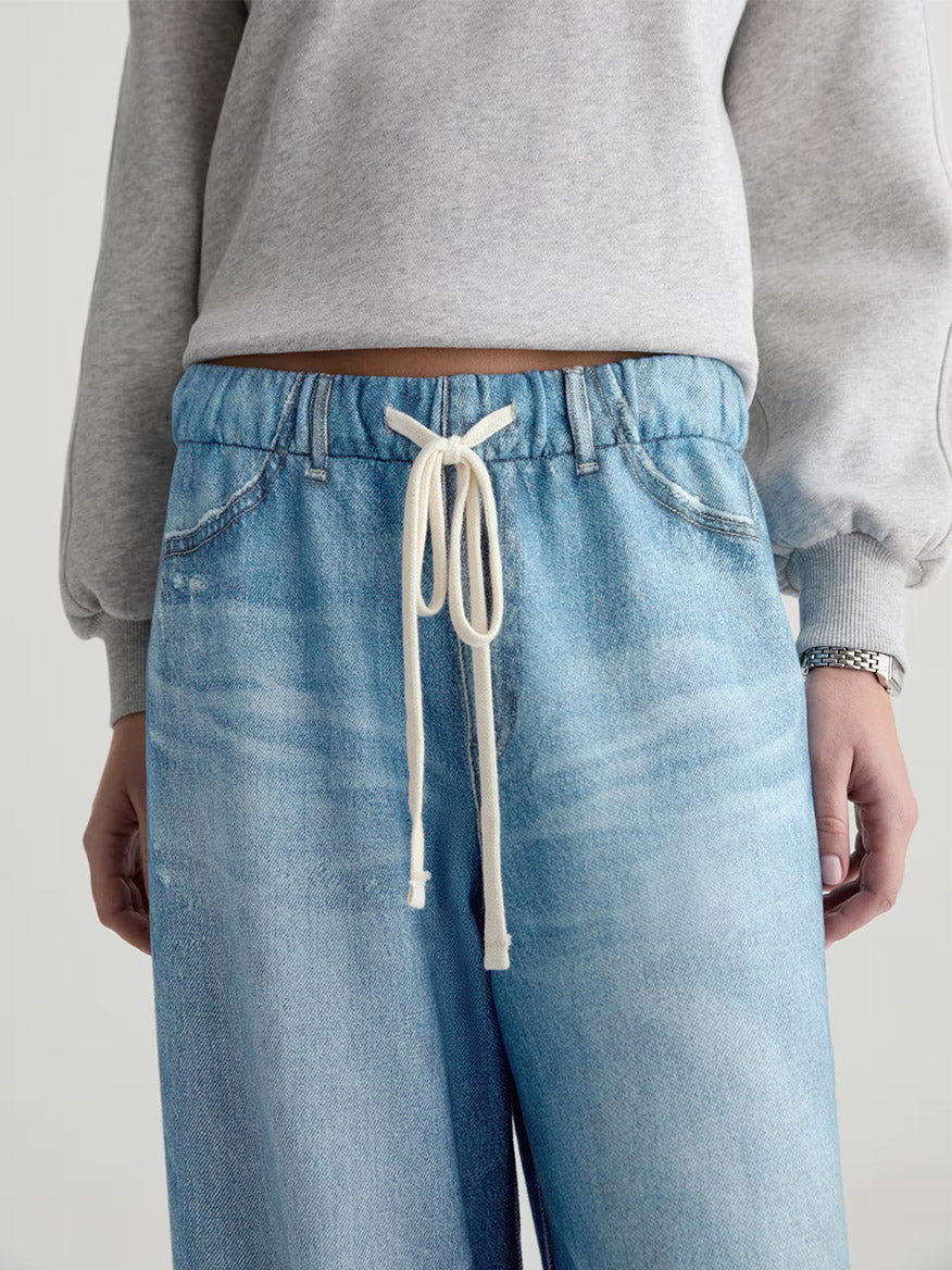 A person wearing AG Jeans Stella Off-Duty Denim Sweatpant in Splash, a light blue French terry style featuring a white drawstring at the waist, paired with a light grey sweatshirt.