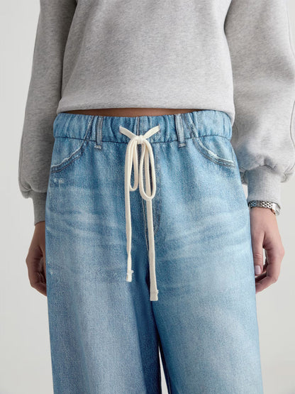 A person wearing AG Jeans Stella Off-Duty Denim Sweatpant in Splash, a light blue French terry style featuring a white drawstring at the waist, paired with a light grey sweatshirt.