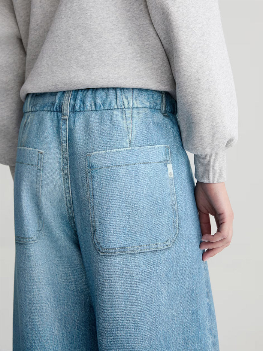 A person wearing AG Jeans Stella Off-Duty Denim Sweatpant in Splash, shown from the waist down with their back to the camera and paired with a light gray French terry sweatshirt.