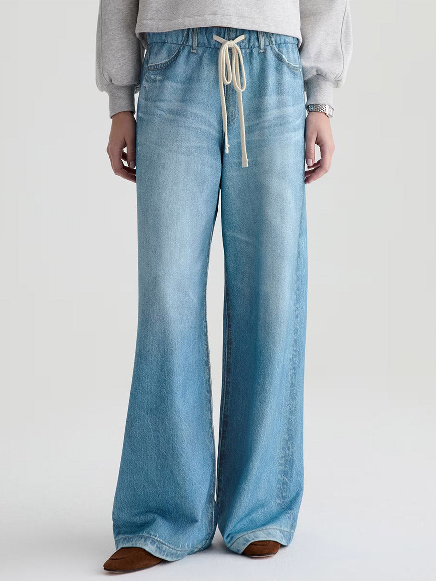 Wearing the AG Jeans Stella Off-Duty Denim Sweatpant in Splash, paired with a grey French terry sweatshirt and brown shoes, this person stands against a plain background.