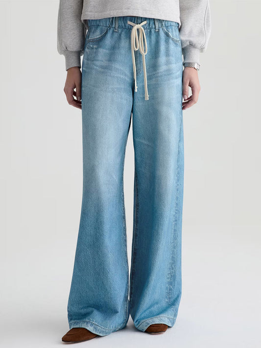 Wearing the AG Jeans Stella Off-Duty Denim Sweatpant in Splash, paired with a grey French terry sweatshirt and brown shoes, this person stands against a plain background.