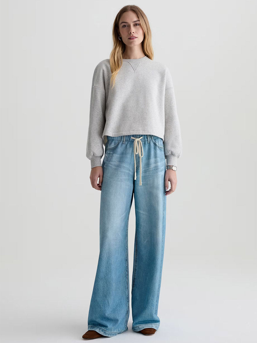 A woman stands against a plain background wearing the AG Jeans Stella Off-Duty Denim Sweatpant in Splash, featuring a light blue wash and a drawstring waist.