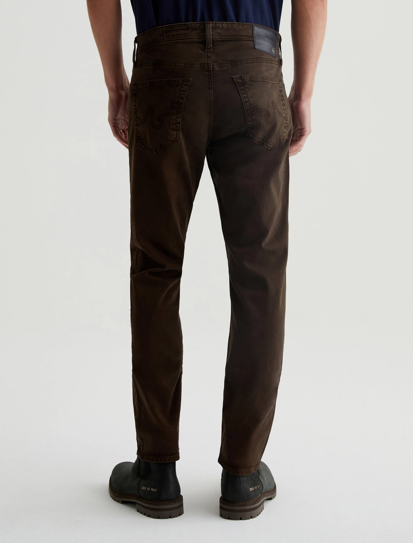 Rear view of a person wearing AG Jeans Tellis Sueded Modern Slim in Sulfur Deep Woodland and black shoes, standing against a plain background.