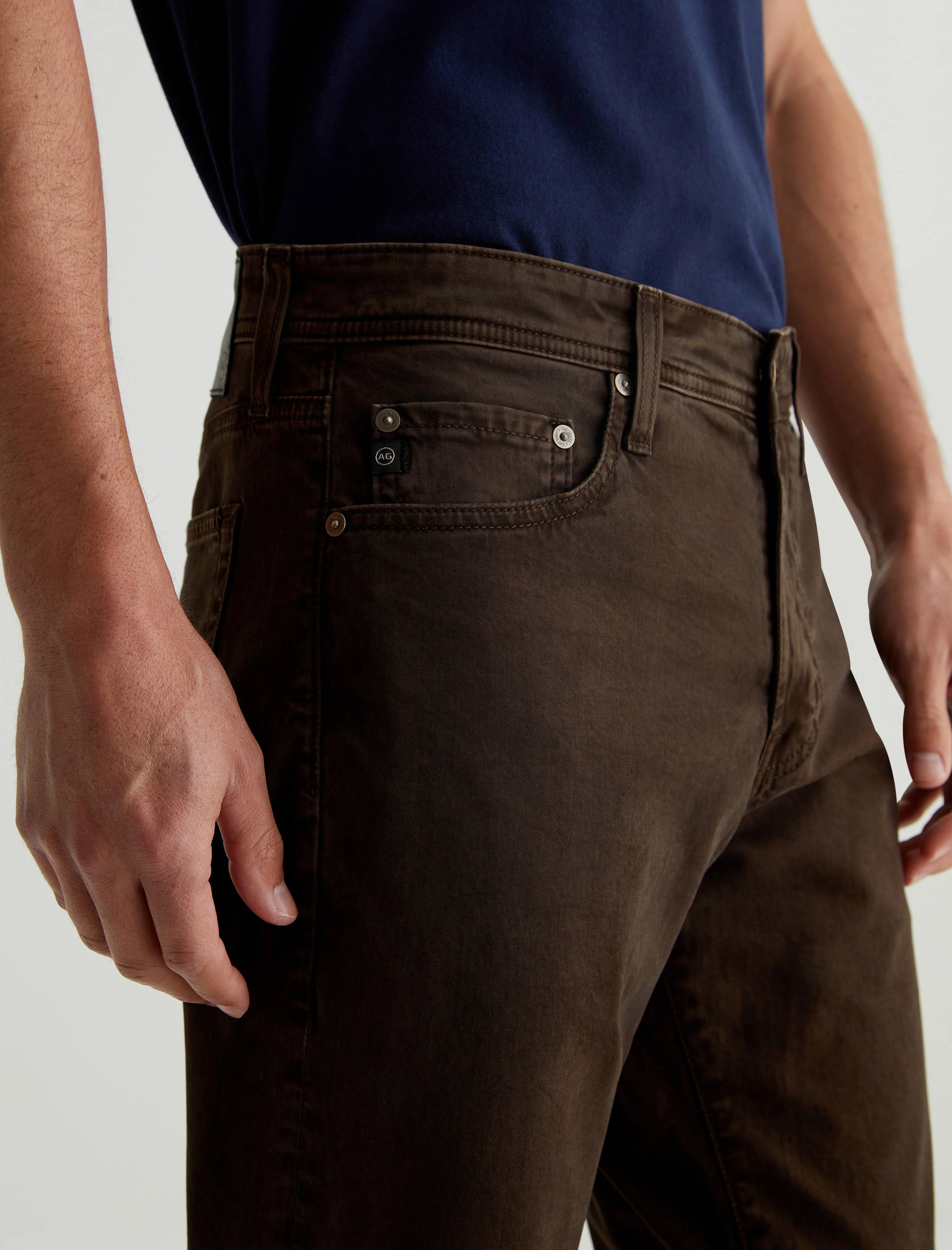 A person is shown from mid-torso to knee, standing against a neutral background, wearing AG Jeans Tellis Sueded Modern Slim pants in Sulfur Deep Woodland.