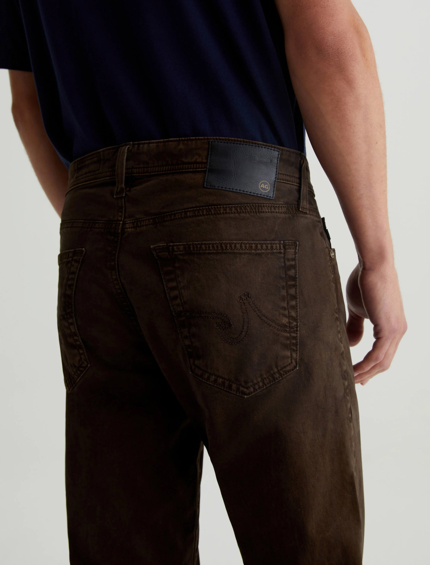 A person wearing the AG Jeans Tellis Sueded Modern Slim in Sulfur Deep Woodland and a navy shirt is shown from behind, highlighting the sueded stretch sateen fabric of the men's pants.