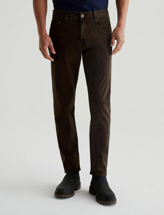 A person stands against a light gray background, showcasing the AG Jeans Tellis Sueded Modern Slim in Sulfur Deep Woodland paired with black boots. The slim fit emphasizes the sueded stretch sateen texture, adding a touch of sophistication.