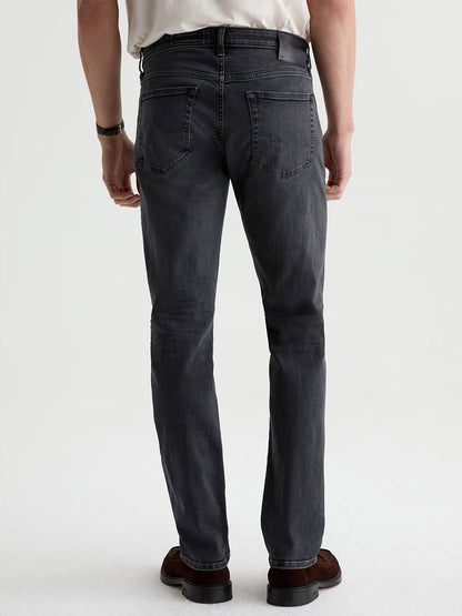 A man stands facing away, wearing AG Jeans Tellis in 7 Years Black Nile, paired with brown shoes and a white shirt. His arms are relaxed at his sides against a plain, light-colored background.