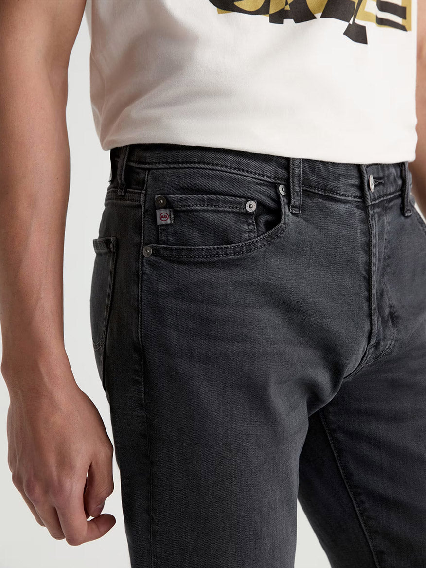 A person wearing a white t-shirt and AG Jeans Tellis in 7 Years Black Nile stands with their left hand relaxed at their side.