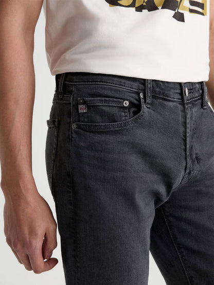 A person wearing a white t-shirt and AG Jeans Tellis in 7 Years Black Nile stands with their left hand relaxed at their side.