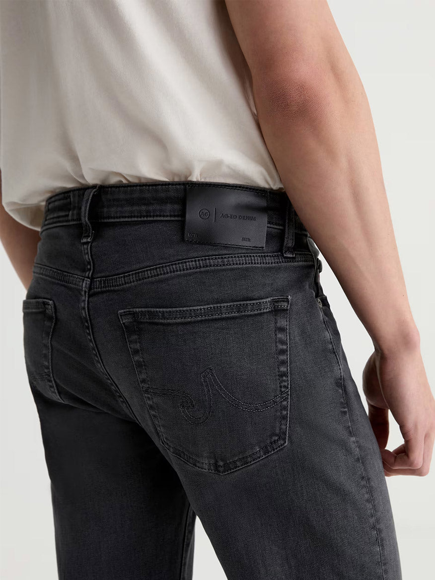 A person wearing the AG Jeans Tellis in 7 Years Black Nile, shown from the back in a white shirt and dark gray jeans, with emphasis on the rear pocket and waistband label. These slim-fit jeans feature a dark grey wash crafted from comfortable stretch denim for a modern silhouette.