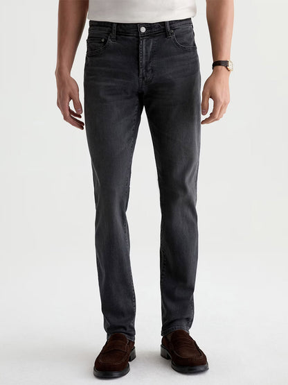 A person wearing a white shirt, AG Jeans Tellis in 7 Years Black Nile (dark grey wash slim jeans made from stretch denim), brown shoes, and a watch stands facing forward with arms relaxed by their sides.