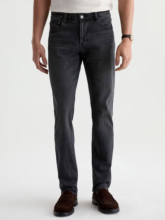 A person wearing a white shirt, AG Jeans Tellis in 7 Years Black Nile (dark grey wash slim jeans made from stretch denim), brown shoes, and a watch stands facing forward with arms relaxed by their sides.