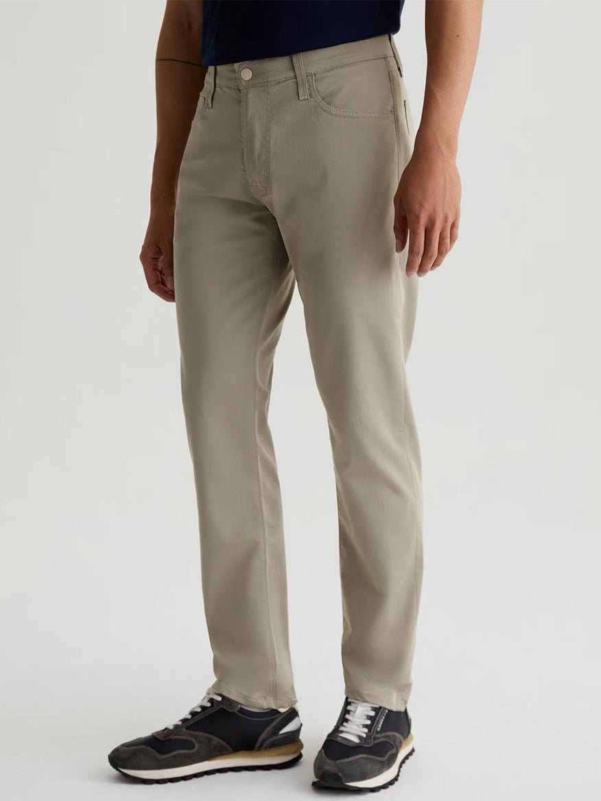 A person wears AG Jeans Tellis Airluxe™ Modern Slim Pant in Dry Dust, paired with a navy shirt and black sneakers, standing against a plain light background.