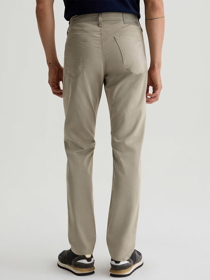 A person is seen from behind wearing AG Jeans Tellis Airluxe™ Modern Slim Pant in Dry Dust, paired with a dark shirt and black sneakers with white soles.