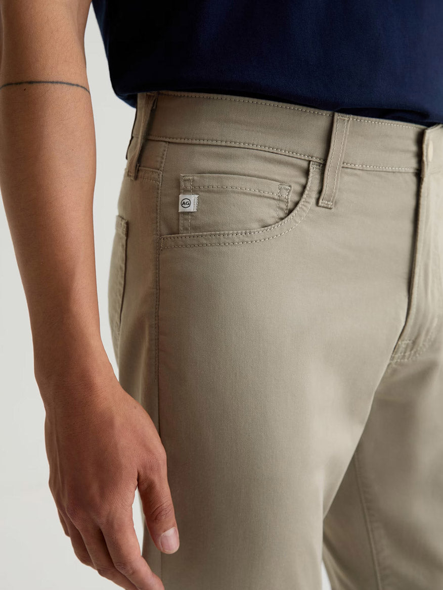 A person wears the AG Jeans Tellis Airluxe™ Modern Slim Pant in Dry Dust with a navy shirt; the image highlights the side pocket and waistband, showing part of their left arm.