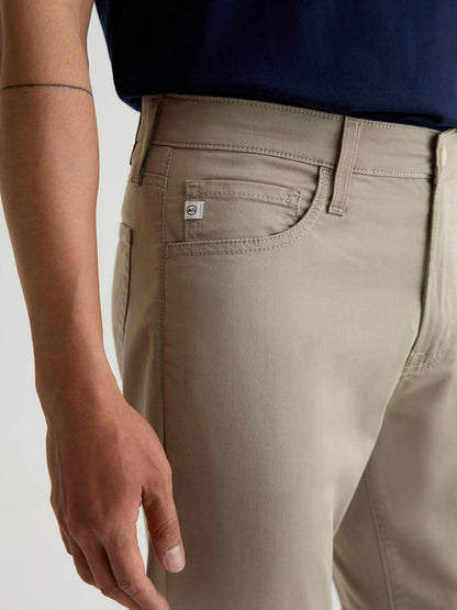 A person wears the AG Jeans Tellis Airluxe™ Modern Slim Pant in Dry Dust with a navy shirt; the image highlights the side pocket and waistband, showing part of their left arm.
