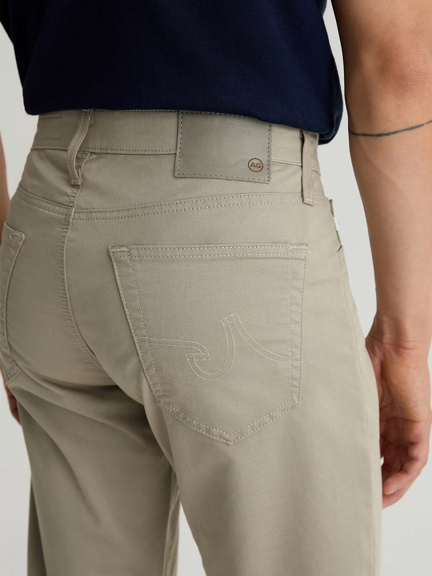 Person wearing AG Jeans Tellis Airluxe™ Modern Slim Pant in Dry Dust, paired with a navy shirt, shown waist down from the back to highlight pocket stitching and a small tattoo on the right arm.