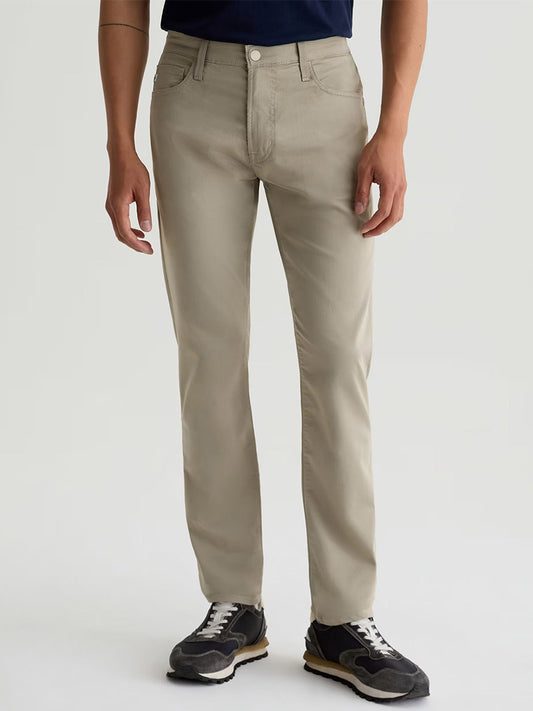 A person wears AG Jeans Tellis Airluxe™ Modern Slim Pant in Dry Dust, a men’s five-pocket slim fit, paired with a dark shirt and black sneakers, standing against a plain light background.