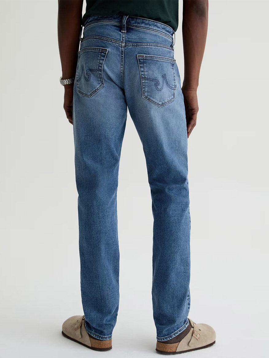 A person stands facing away, wearing AG Jeans Tellis in Horsetail—modern slim jeans in a dark blue wash—paired with a dark shirt, beige shoes, and a watch; shown from the waist down against a plain background.