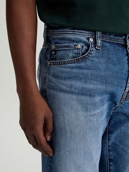 A person wearing AG Jeans Tellis in Horsetail, styled with a dark green top and their left hand resting by their side.