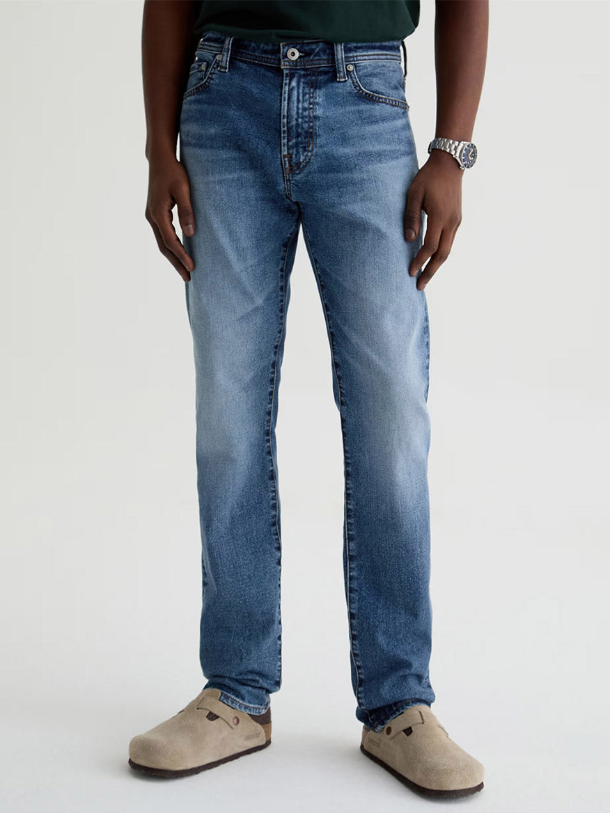 A person stands against a plain light background wearing AG Jeans Tellis in Horsetail, a dark green shirt, beige slip-on shoes, and a silver wristwatch.