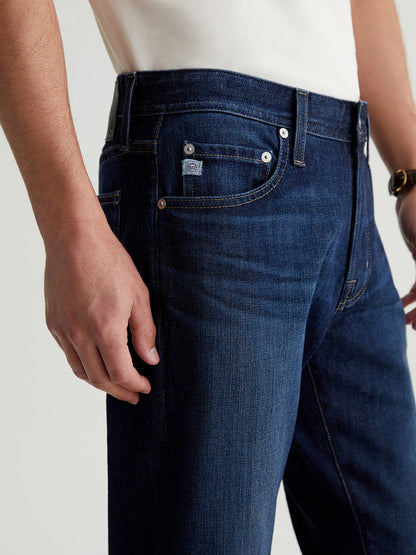 A person is wearing the AG Jeans Tellis in Piazza, featuring a sleek design and flexibility with its dark indigo wash, paired with a white shirt. The image emphasizes the hand and pocket area, highlighting the comfort of Flex 360° Denim.