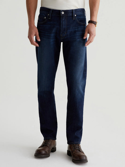 A person is wearing the AG Jeans Tellis in Piazza, slim men's jeans in a dark indigo wash, paired with a white shirt, brown leather shoes, and a watch.