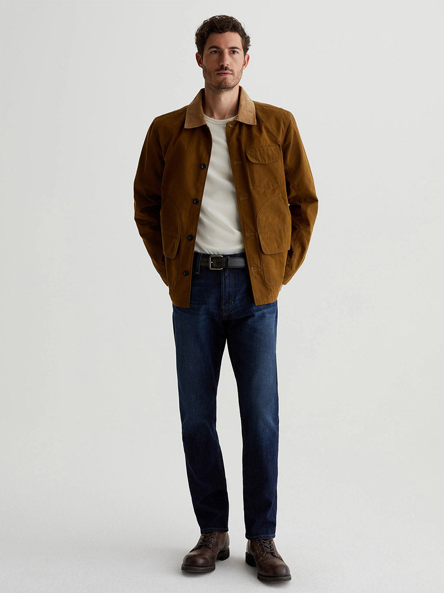 A man stands wearing a brown jacket and white shirt, paired with AG Jeans Tellis in Piazza, featuring a dark indigo wash for his slim men's jeans. His look is completed with brown shoes against a plain background.