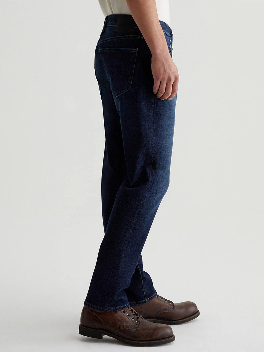 A person stands sideways against a plain background, wearing the AG Jeans Tellis in Piazza—a pair of slim men's jeans in dark indigo wash—paired with brown shoes, highlighting the versatility of Flex 360° Denim.