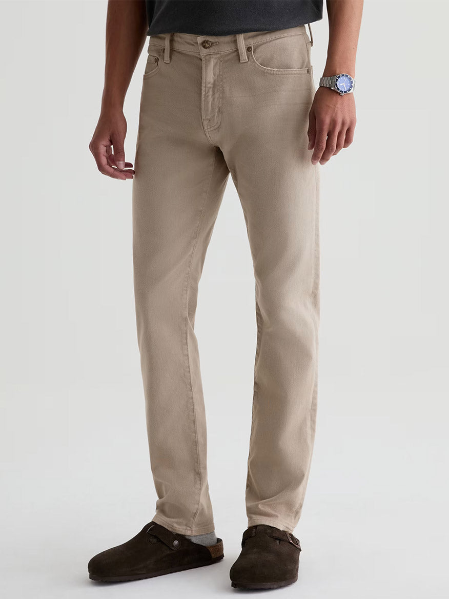 A person wears AG Jeans Tellis in 7 Years Sulfur Stone Barrack, a dark shirt, brown suede clogs, and a wristwatch while standing against a plain light background.