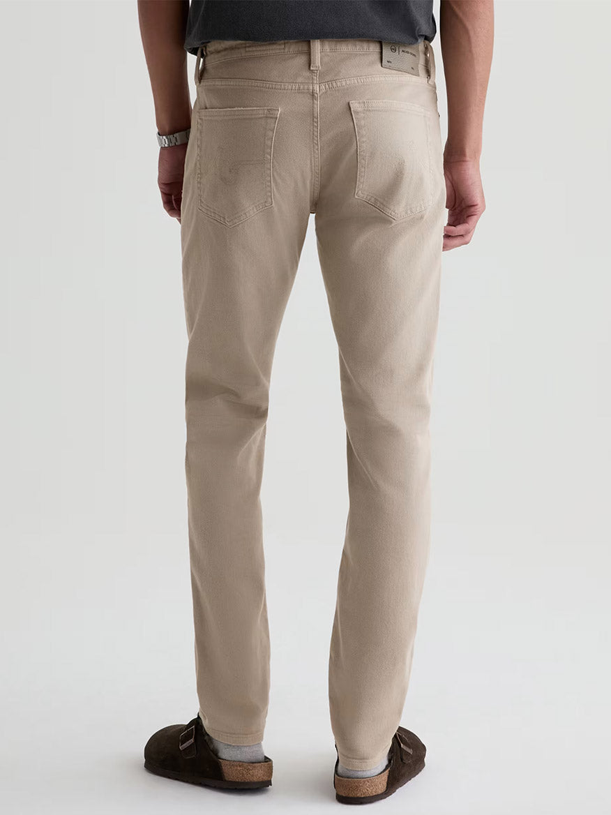 A person in AG Jeans Tellis in 7 Years Sulfur Stone Barrack, paired with a dark shirt, watch, and brown shoes, stands facing away to showcase the stylish back view of these men’s jeans.