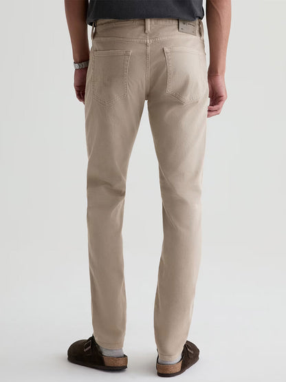 A person in AG Jeans Tellis in 7 Years Sulfur Stone Barrack, paired with a dark shirt, watch, and brown shoes, stands facing away to showcase the stylish back view of these men’s jeans.