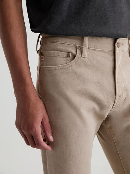 A person wearing a black shirt and beige men's jeans, shown from mid-torso to thigh with one arm relaxed, showcases the AG Jeans Tellis in 7 Years Sulfur Stone Barrack—a stylish and comfortable choice for a refined casual look.