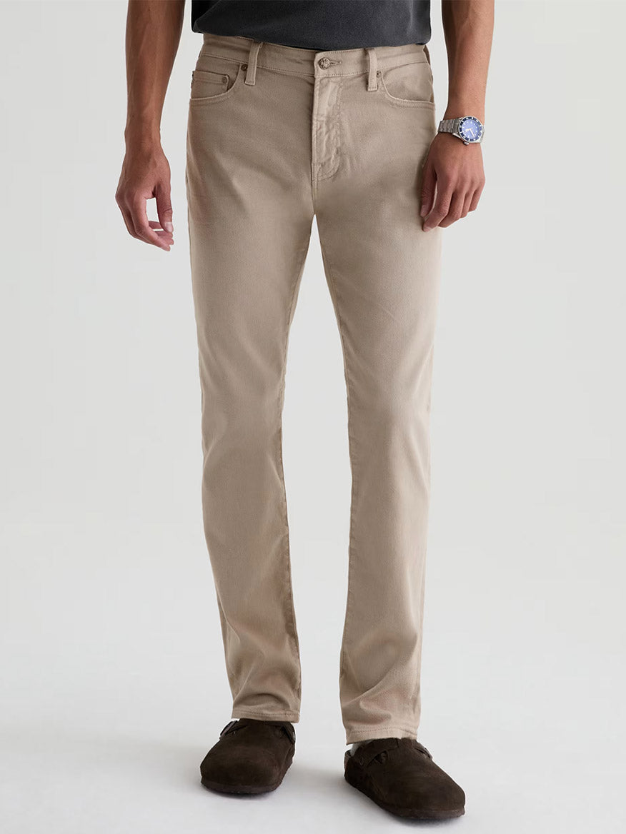 A person in AG Jeans Tellis in 7 Years Sulfur Stone Barrack, a dark shirt, brown shoes, and a wristwatch stands against a plain light background.