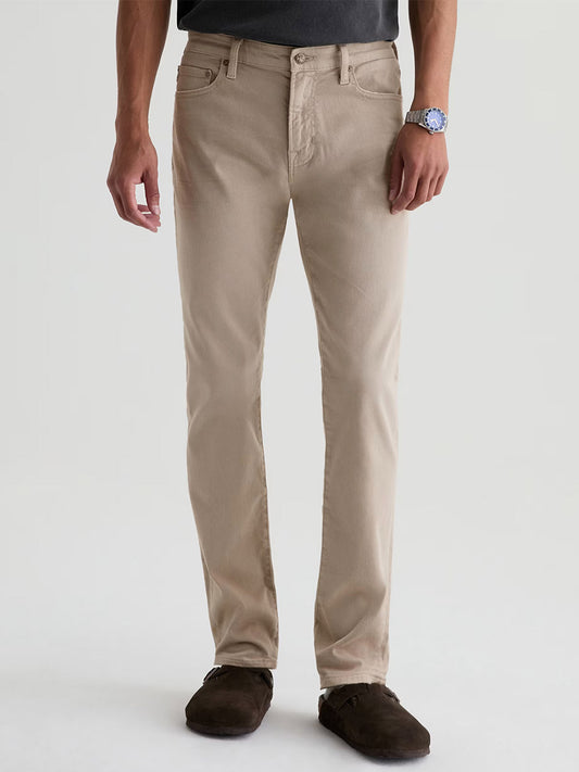 A person in AG Jeans Tellis in 7 Years Sulfur Stone Barrack, a dark shirt, brown shoes, and a wristwatch stands against a plain light background.